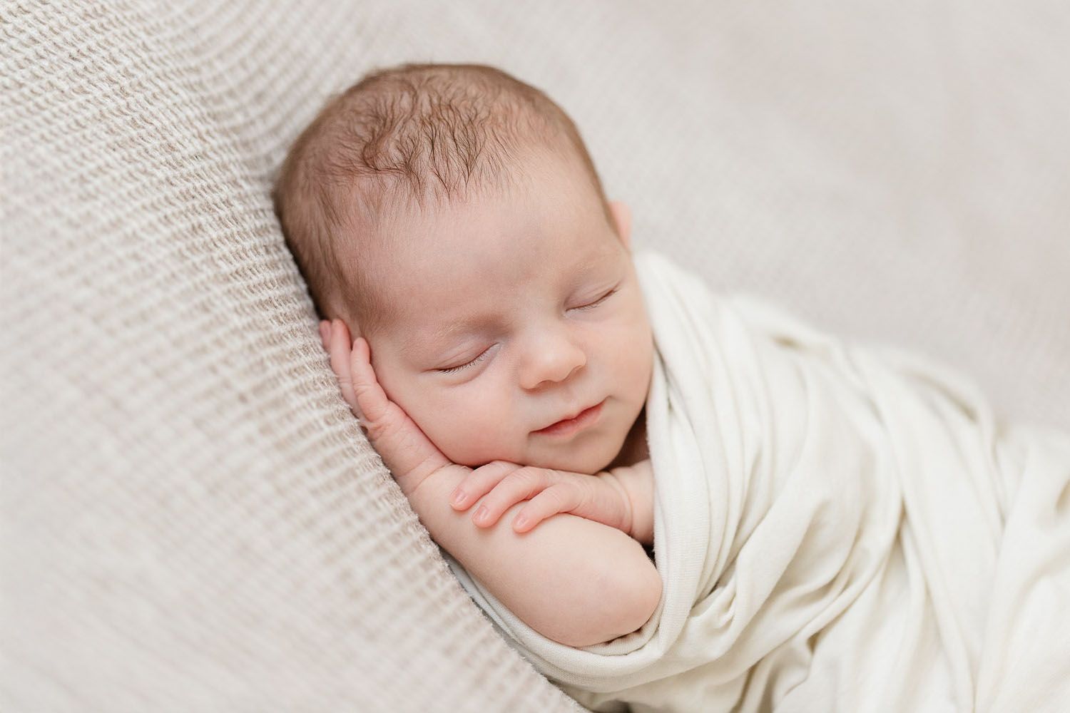 Ein schlafendes, neugeborenes Baby, eingewickelt in weichen weißen Stoff, ruht friedlich auf einem grauen, strukturierten Hintergrund.