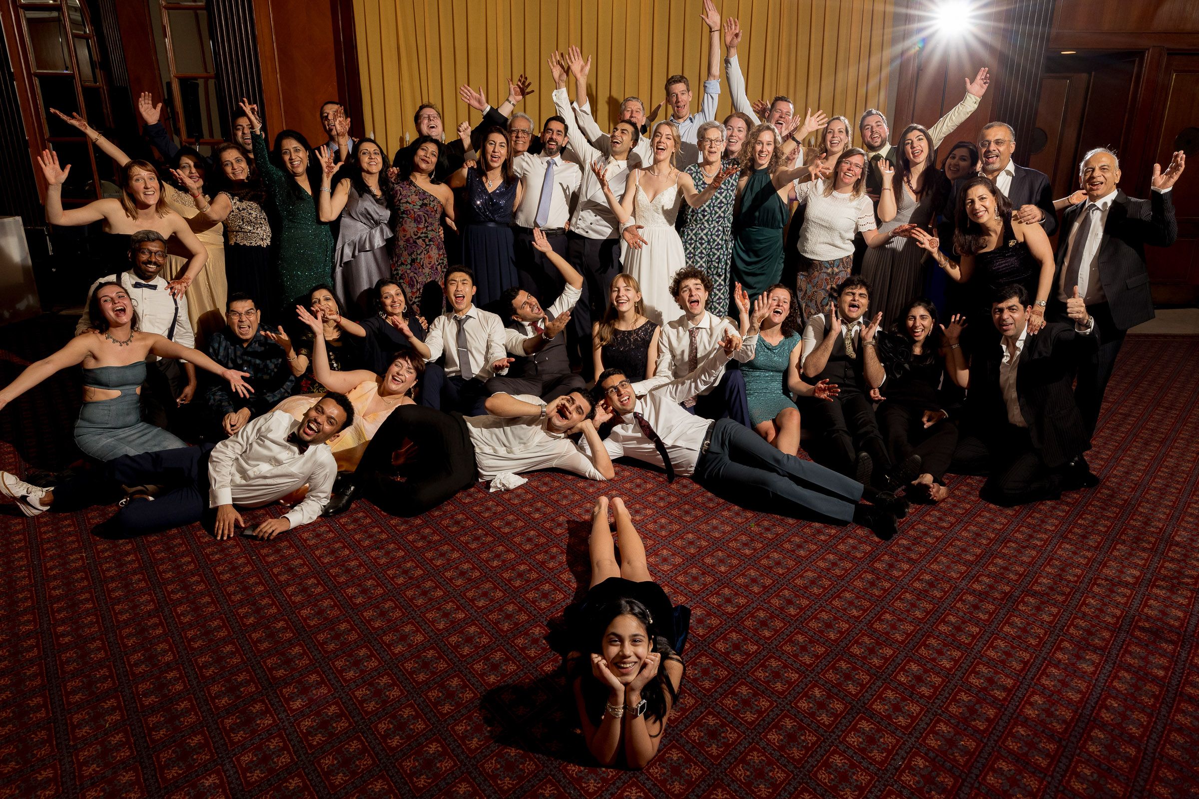 Wedding guests lay on red carpet dance floor while photographer captures unique party shot from above at reception.