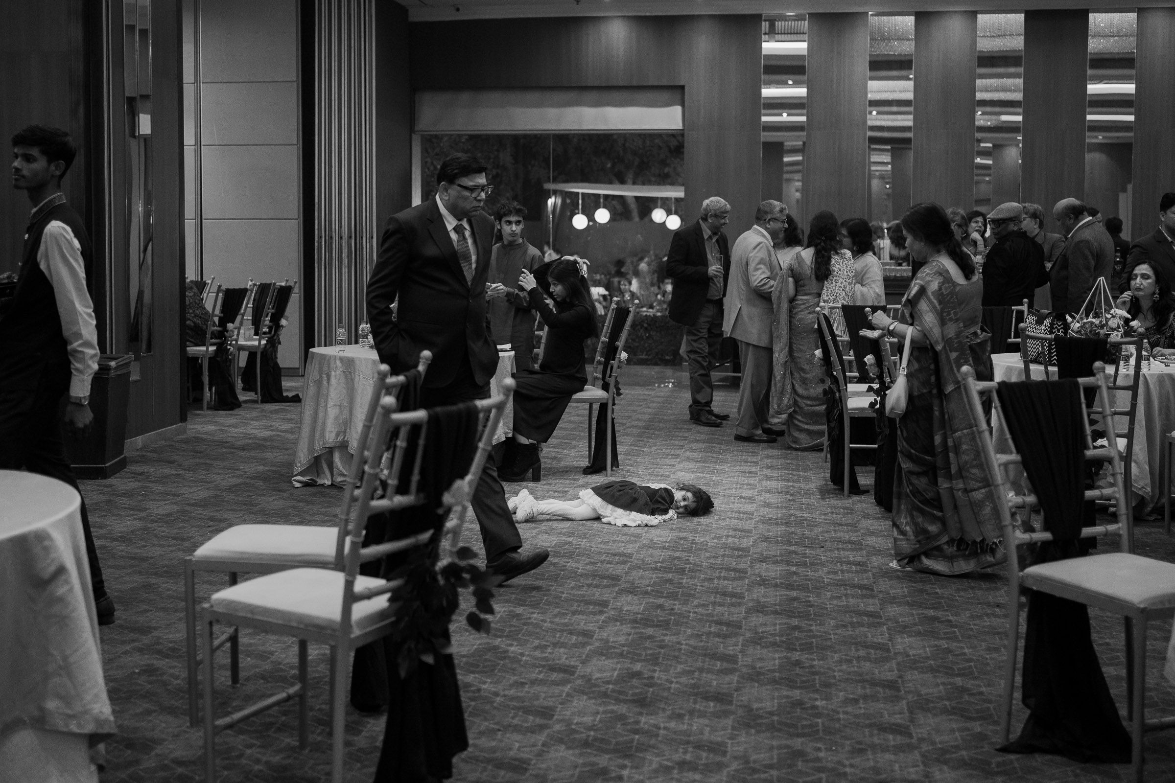 A black and white scene of a crowded dining room or restaurant space with people mingling and tables set up.