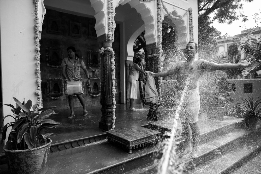 Black and white photograph showing a person being sprayed with water from a hose on a covered porch with ornate columns.