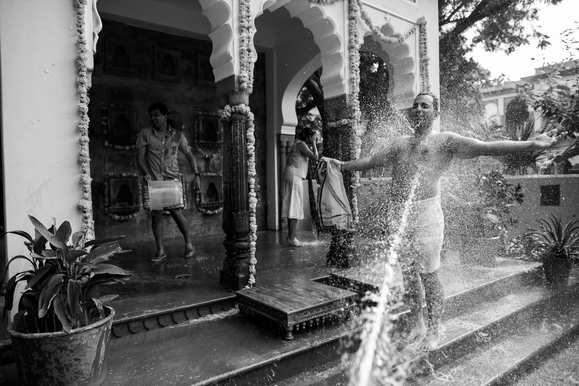 Black and white photograph showing a person being sprayed with water from a hose on a covered porch with ornate columns.
