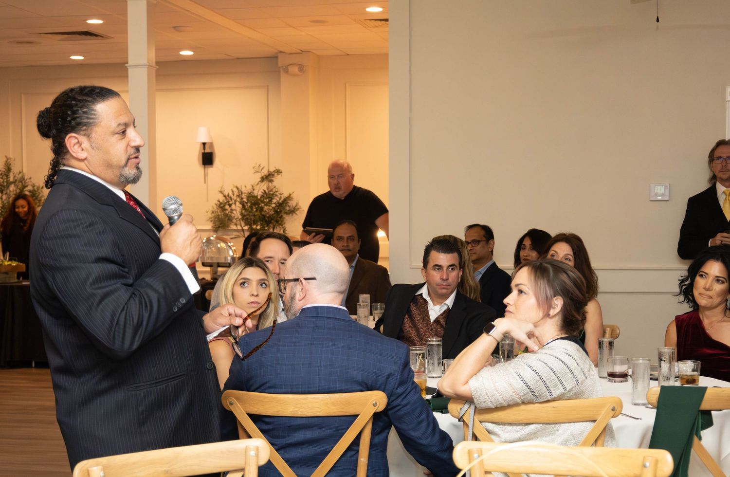 A man in a suit speaks to a seated audience at a formal dinner event in a well-lit banquet hall.