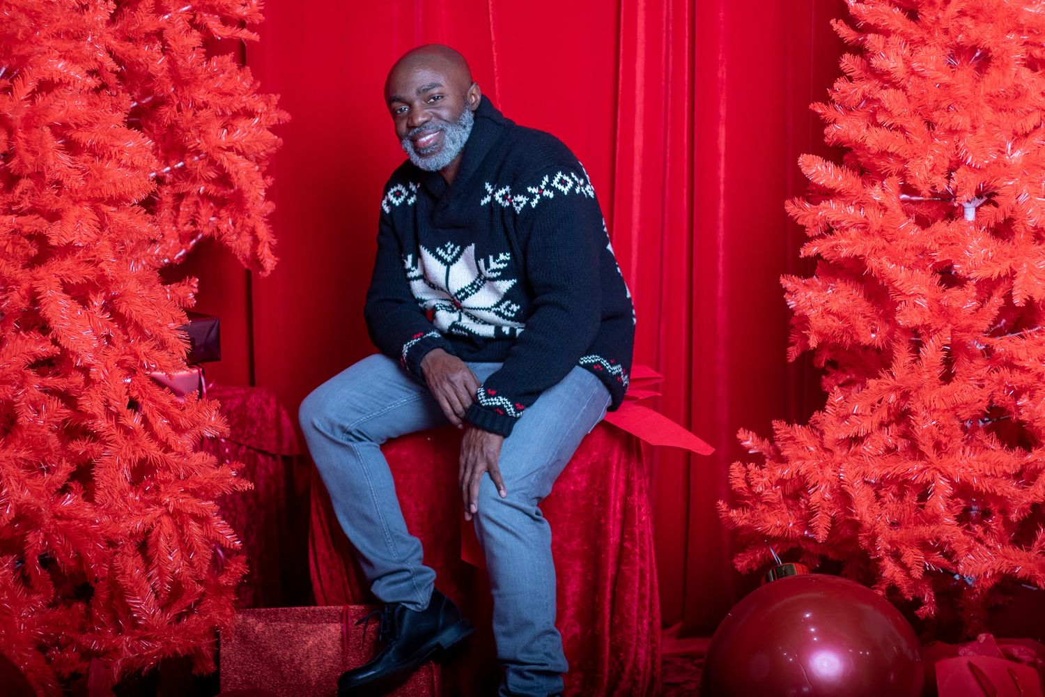Person wearing black holiday sweater seated between bright red artificial Christmas trees.