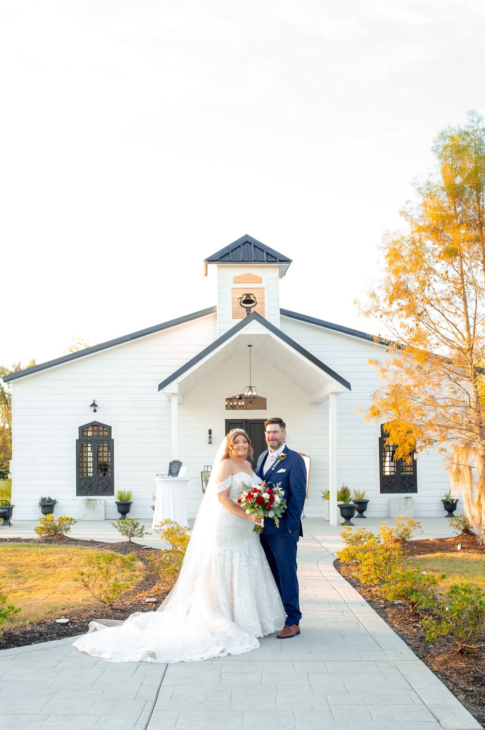 Newlyweds pose in front of a charming white chapel with fall colors in the background.