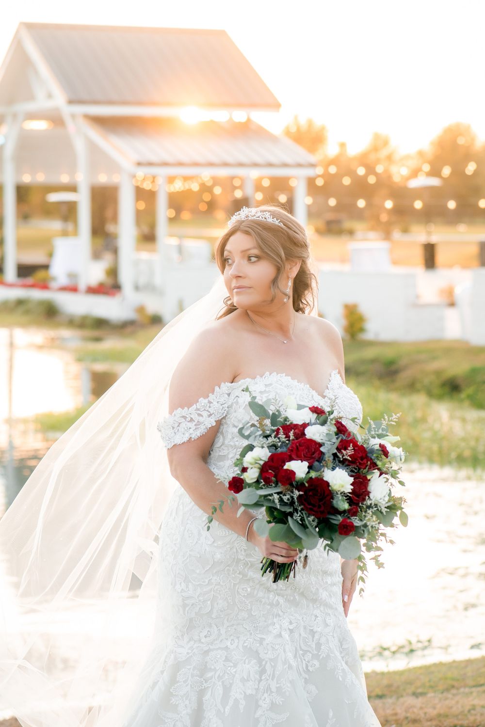 Bride with flowing veil holds red and white bouquet during golden hour.