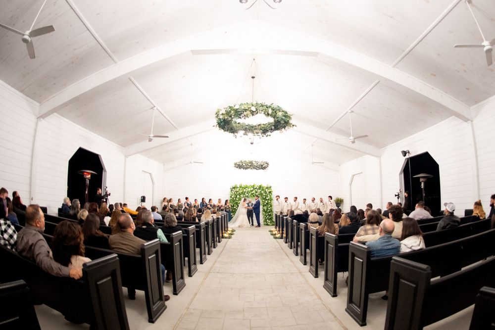 Wedding ceremony venue with rows of white chairs and floral arch decorations in a bright white room.