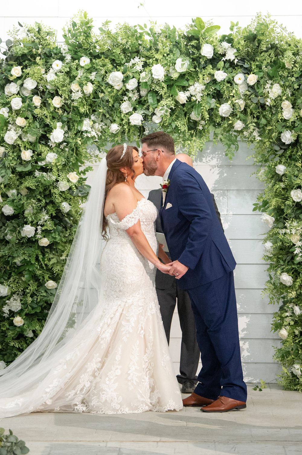 Wedding ceremony moment captured under a lush white rose and greenery arch.