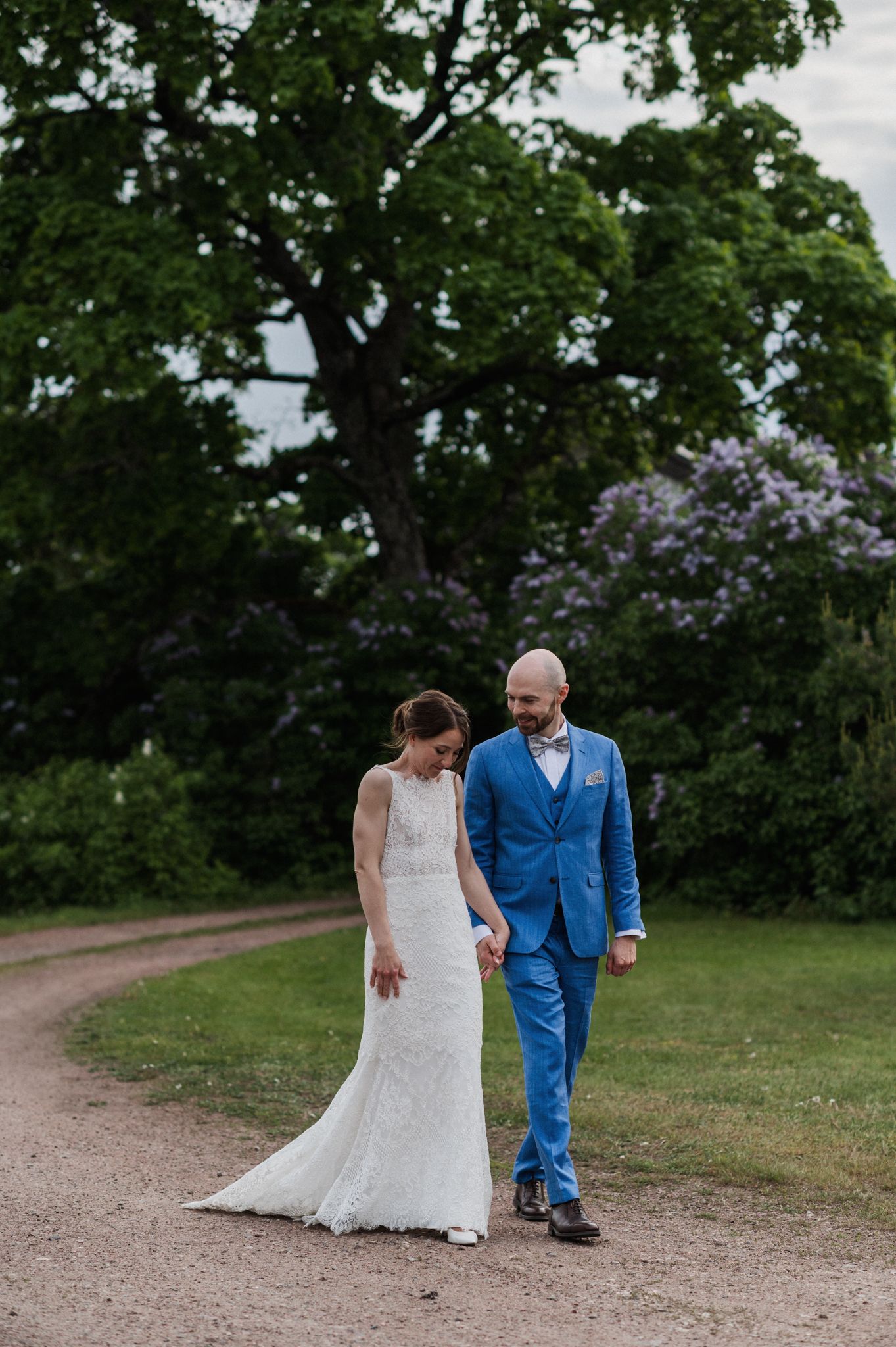 A couple walks along a garden path, with the bride in white lace and the groom wearing a vibrant blue suit.