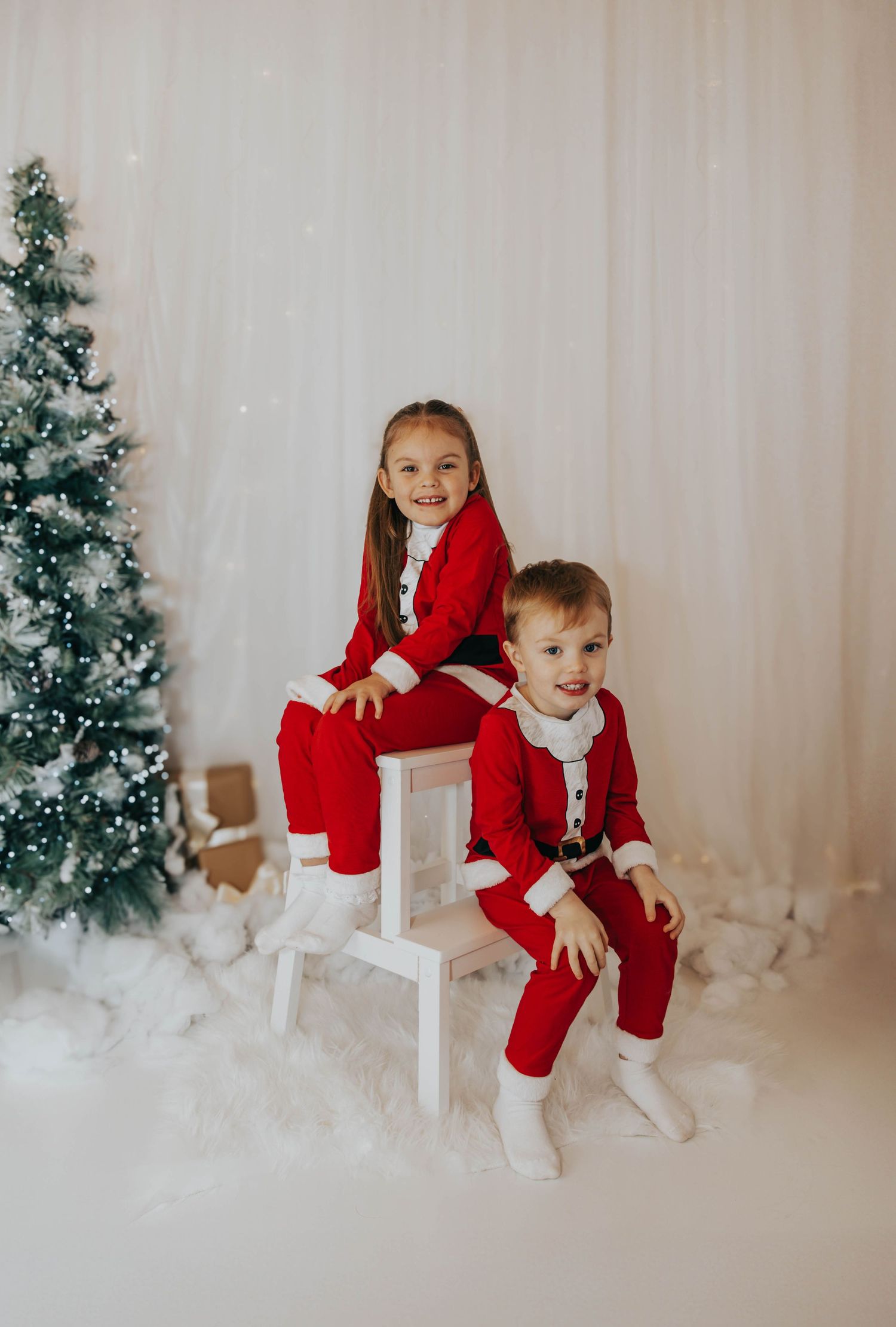 Siblings in matching red and white outfits pose on white steps near Christmas tree.