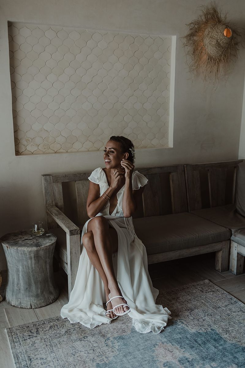 Bride in white dress putting on earrings while seated on a wooden bench in an elegant boho-styled room.