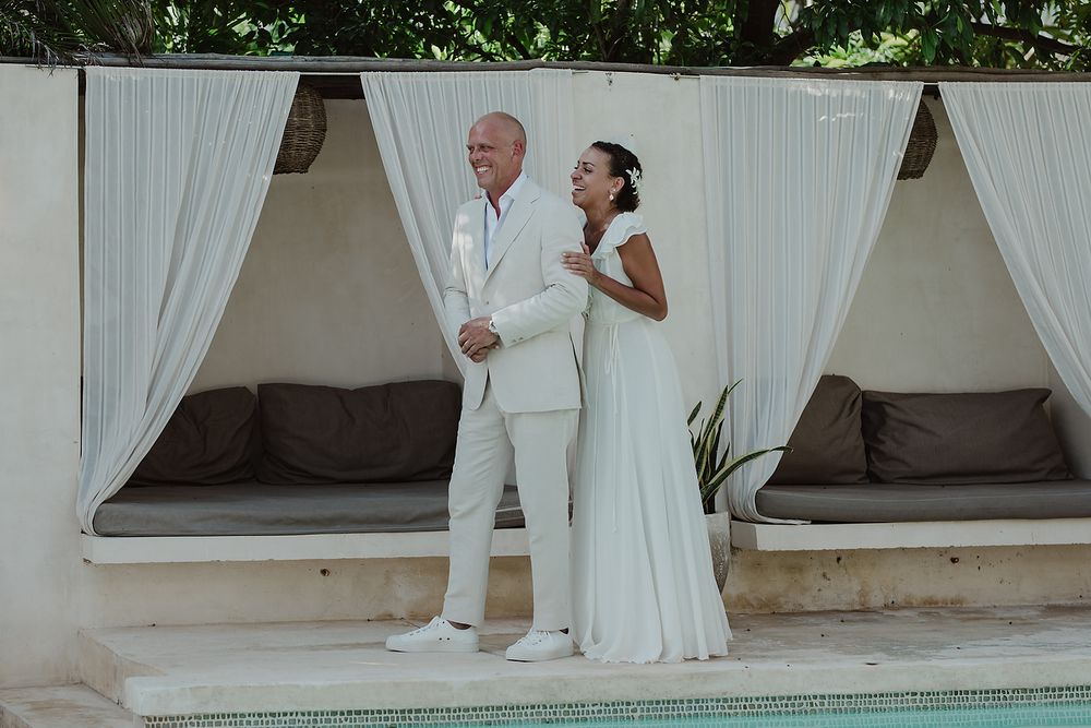 Bride and groom in matching white outfits standing back-to-back by a pool with white draped cabanas.