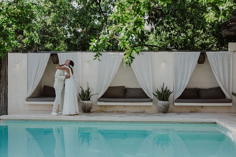Newlyweds sharing a romantic embrace by a serene outdoor pool surrounded by lush greenery and cabanas.