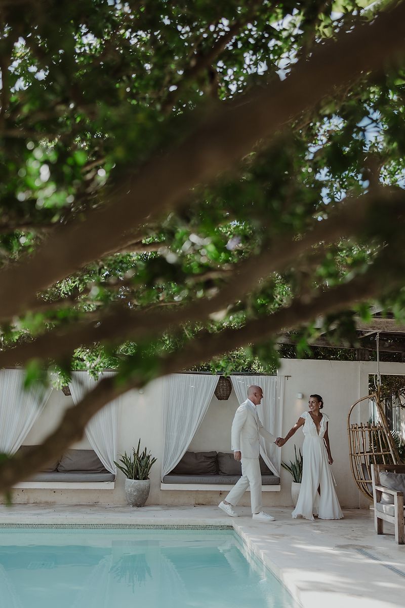 Couple in white wedding attire holding hands by a pool, framed by lush green tree branches at a tropical venue.