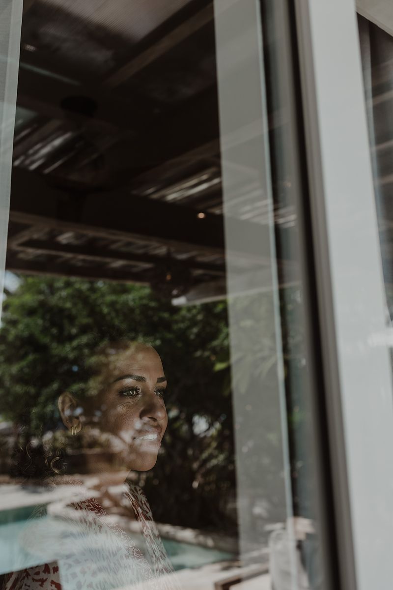 Woman gazing thoughtfully through a glass window with green trees reflected in the pane.