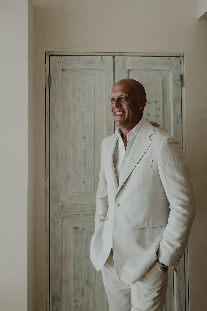 Smiling bald man in a white linen suit standing confidently by a rustic wooden door.