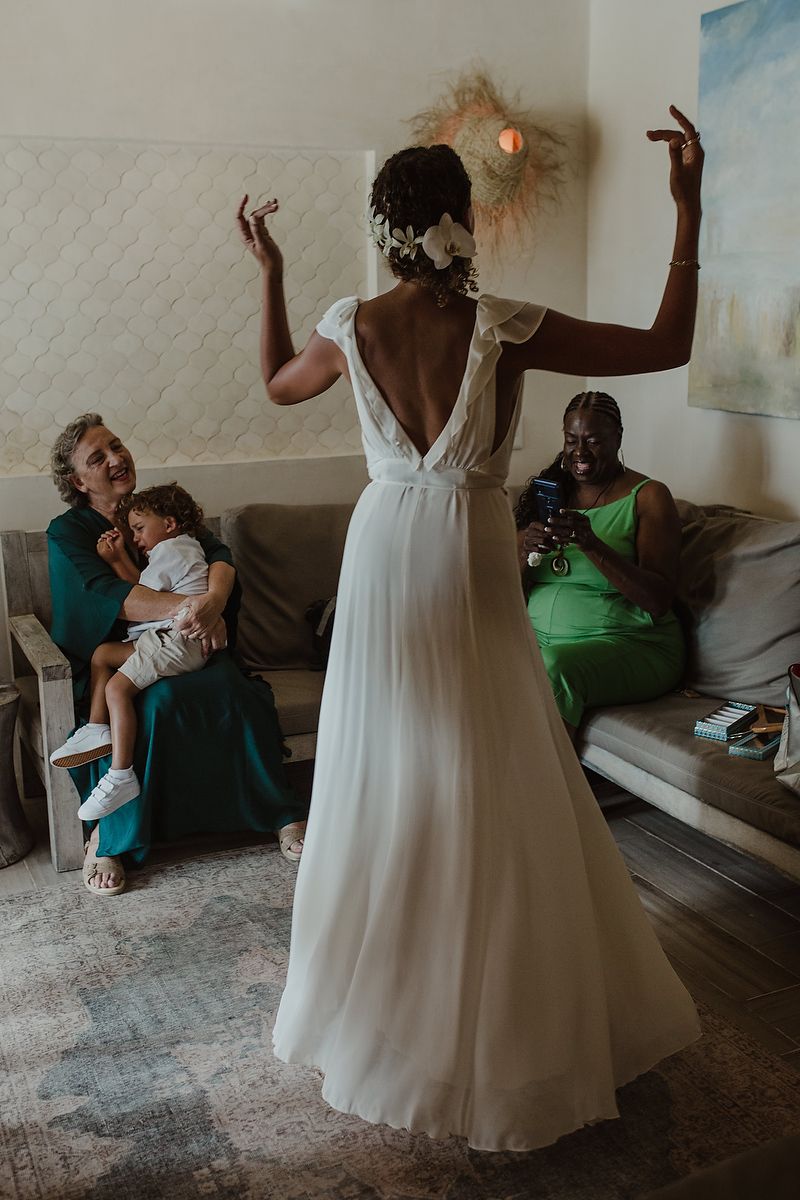 Bride in white backless wedding gown dancing joyfully while family watches and smiles in a cozy room.