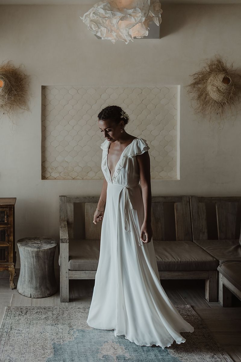 Bride in a flowing white V-neck wedding gown with flutter sleeves stands in a bohemian-styled room.