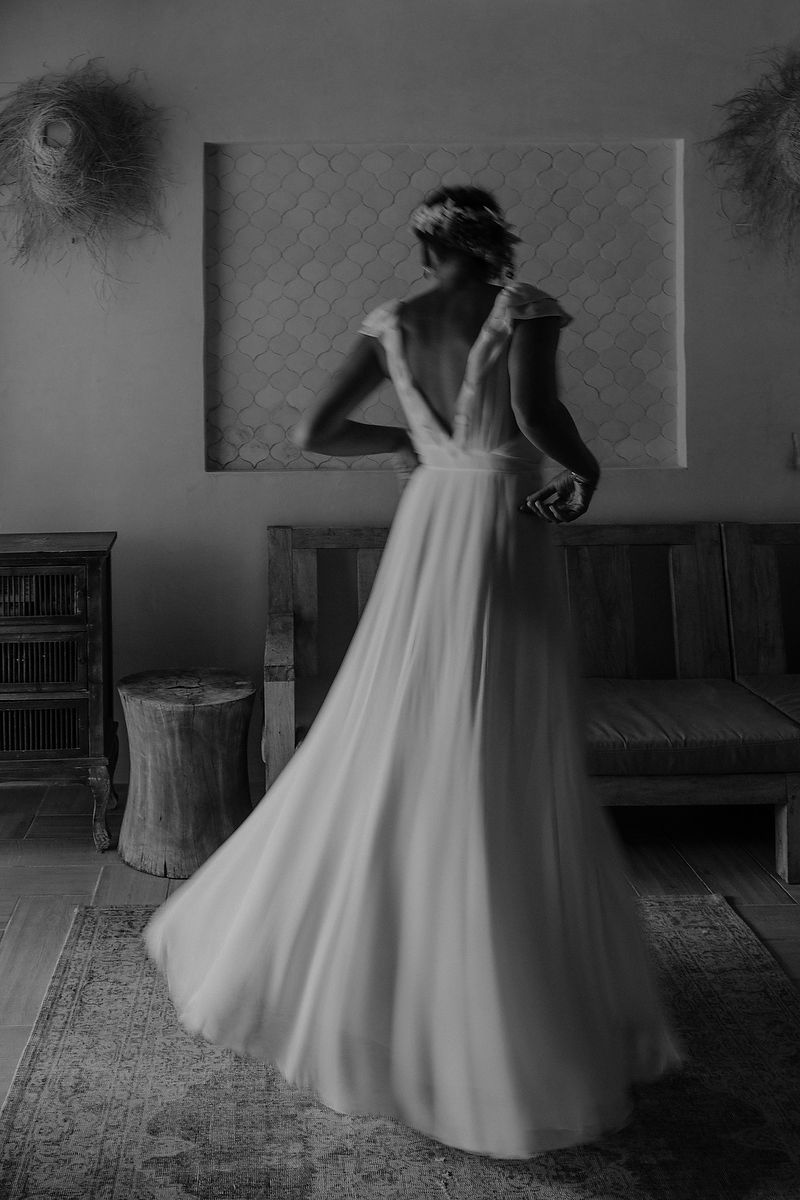 Elegant bride in a flowing white backless wedding dress poses gracefully in a dimly lit room.