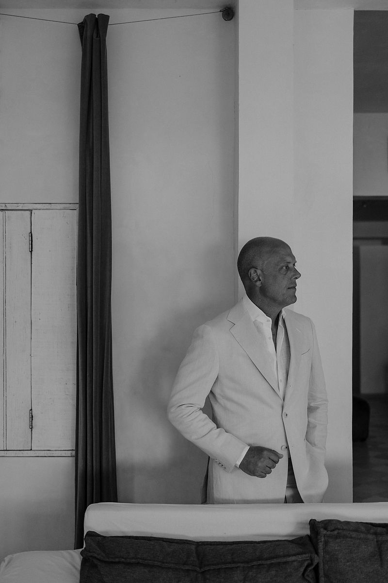 Bald man in white suit standing thoughtfully by dark curtains in black and white portrait photo.