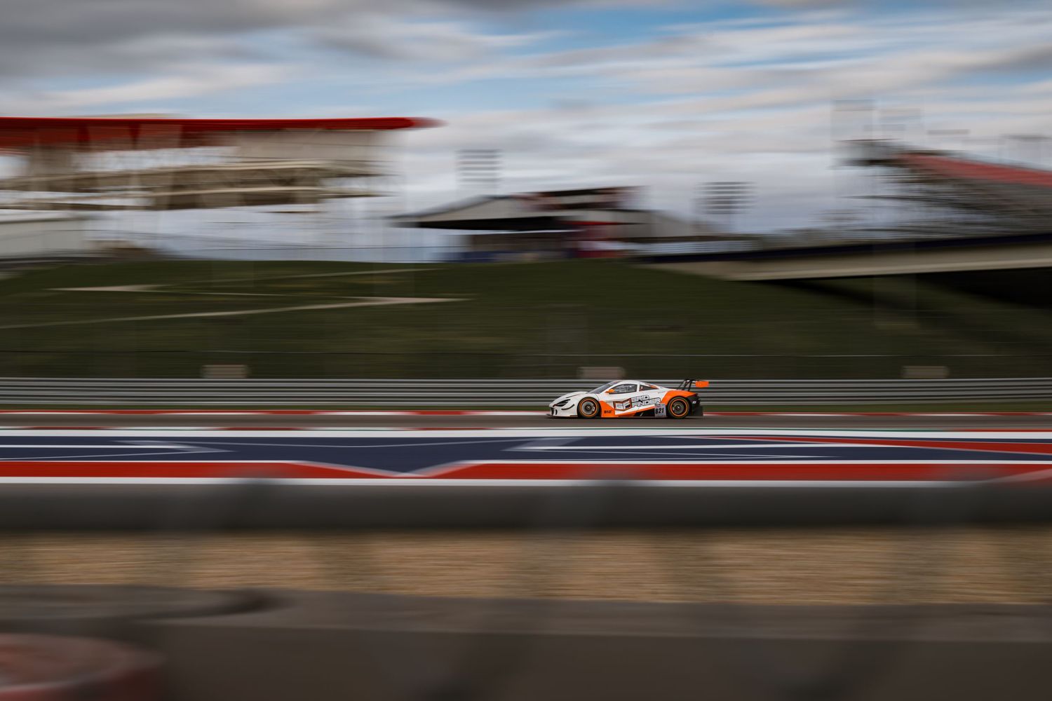 Racing car speeds along a racetrack with motion blur against cloudy sky and mountains in the background.