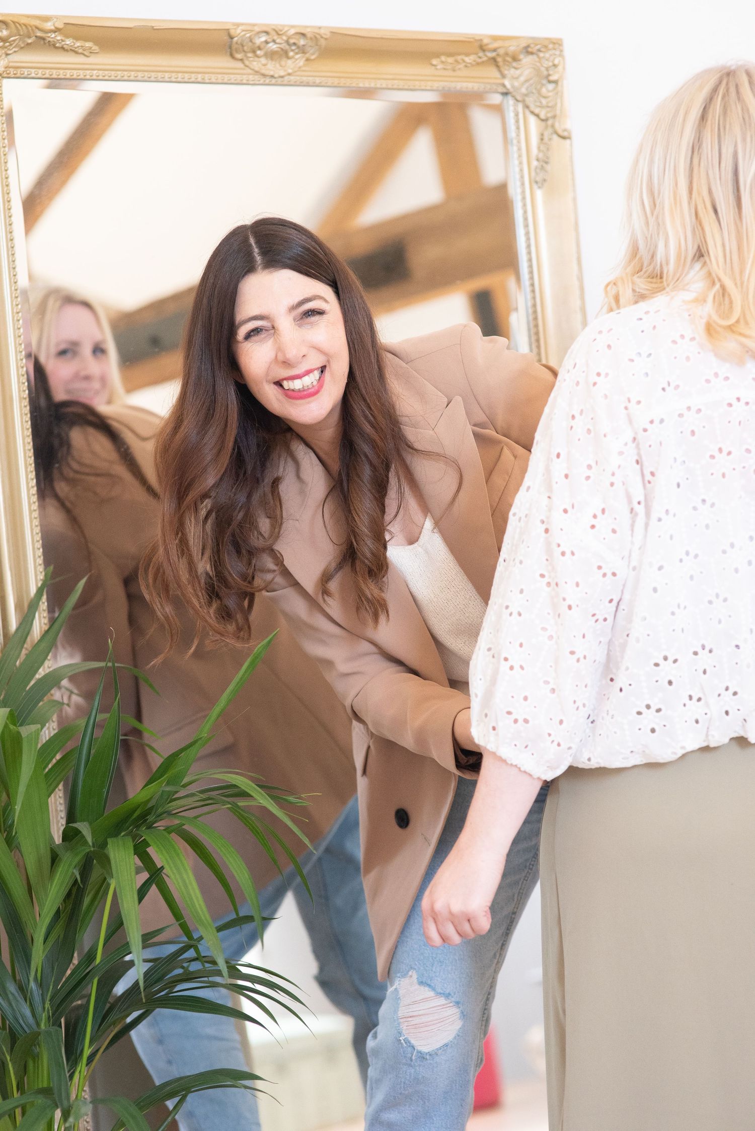 A person in a tan coat smiles while looking through a decorative gold frame with a houseplant in the foreground.