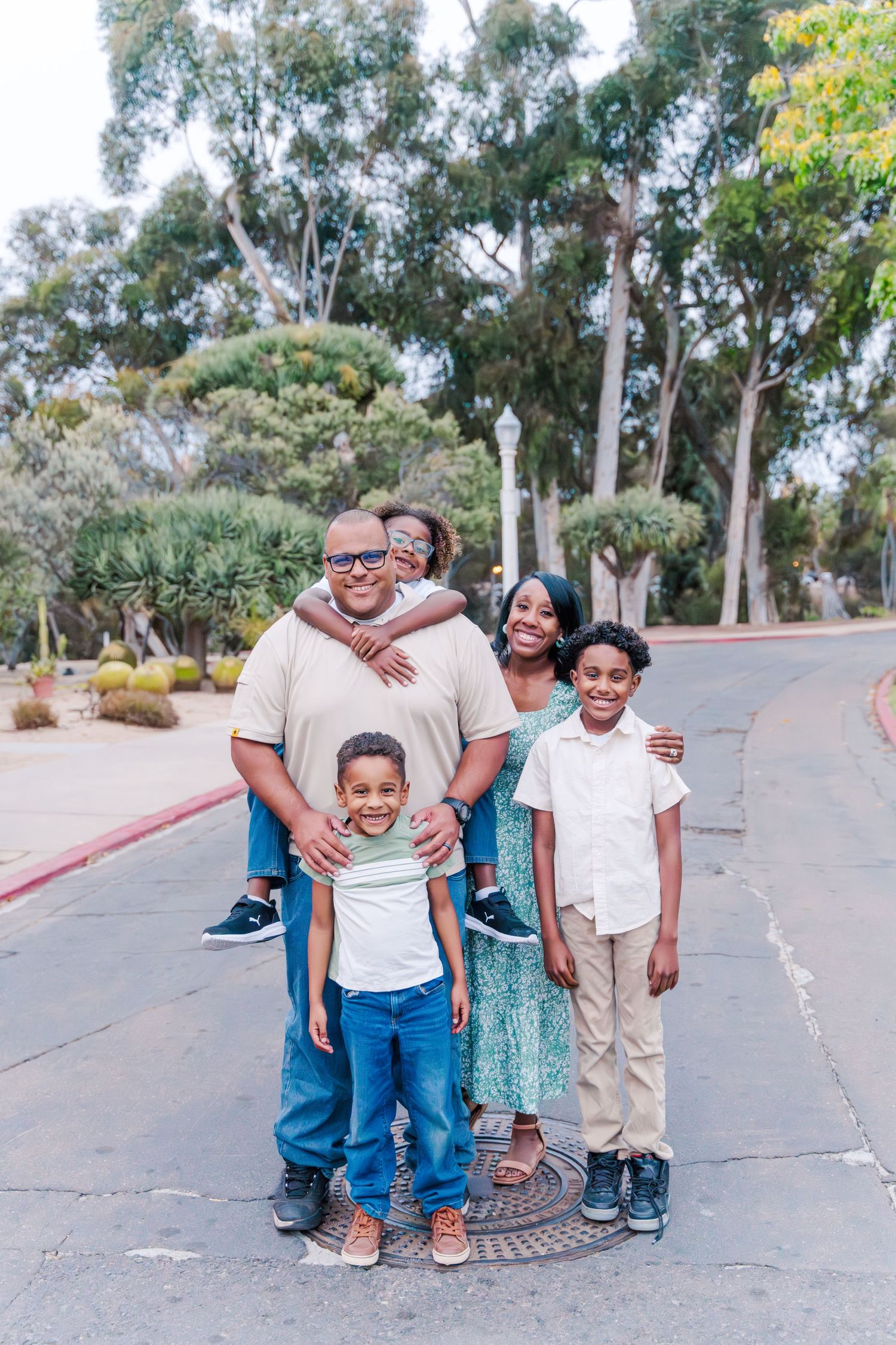 A happy family stands together on a paved path with eucalyptus trees in the background on a sunny day.