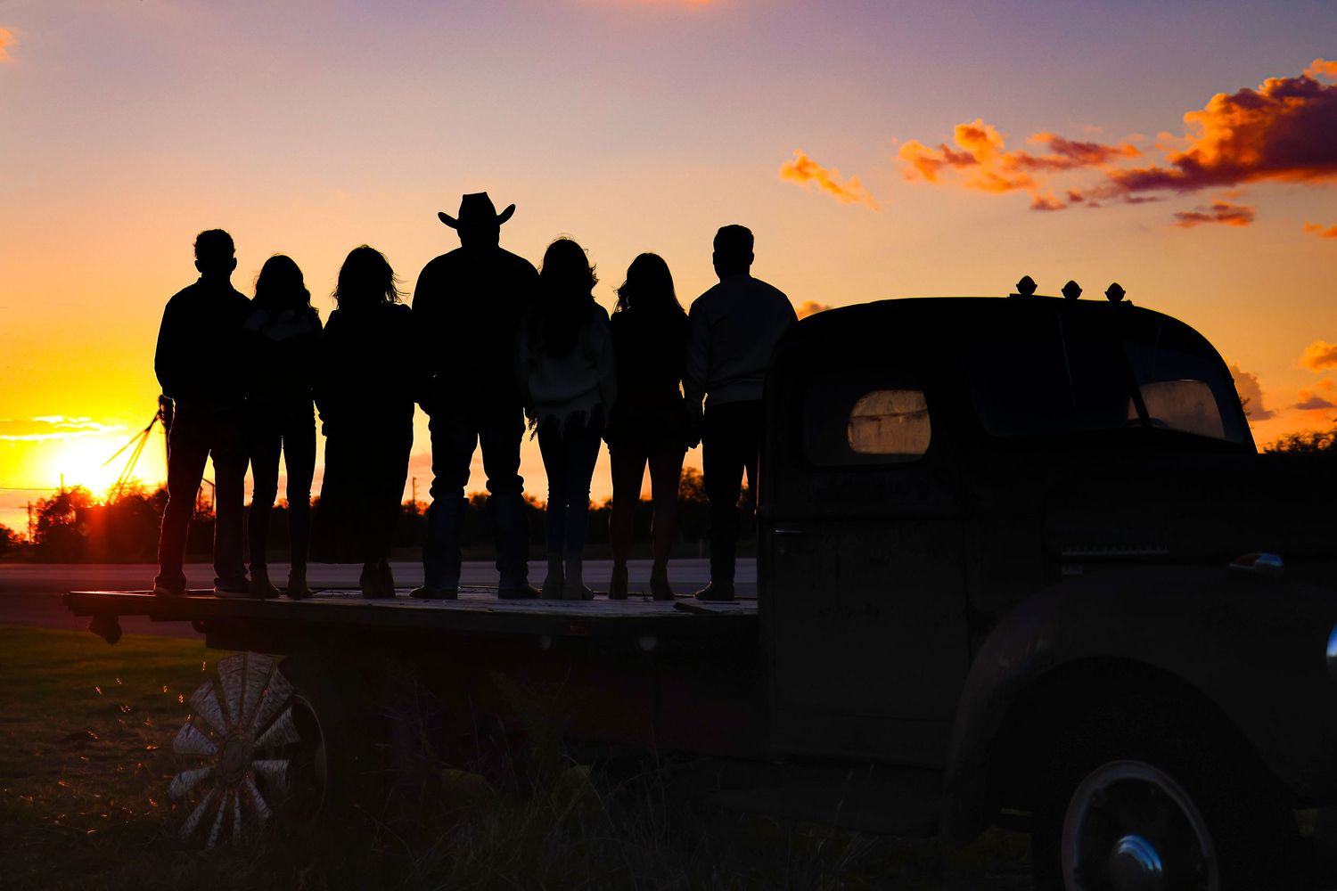 Group of silhouettes standing on a vintage truck against an orange sunset.