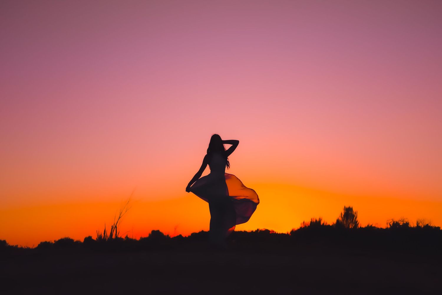 Silhouette of figure in flowing dress against vibrant orange and pink sunset sky.