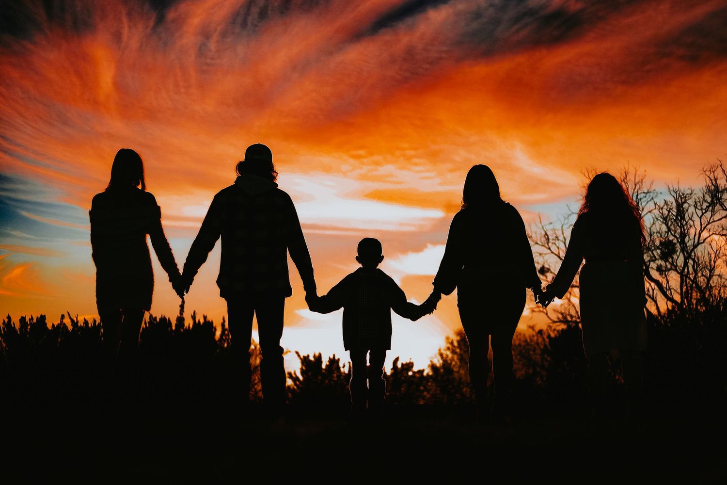 Silhouette of family holding hands against fiery orange sunset sky.