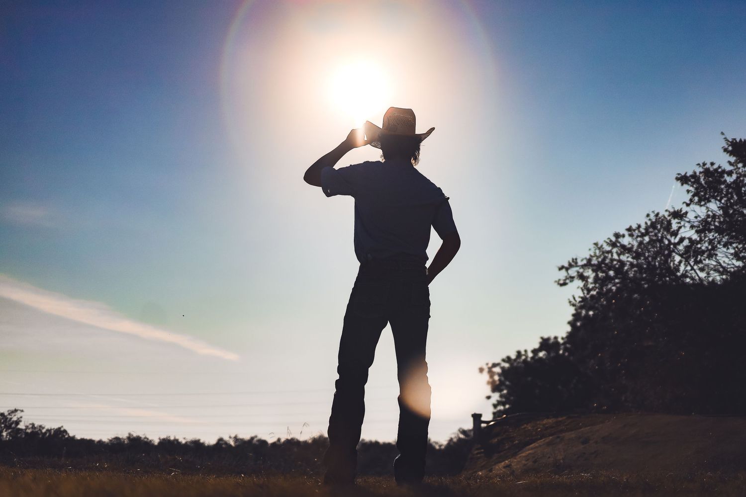 Silhouette of person in cowboy hat looking at sun against blue sky.