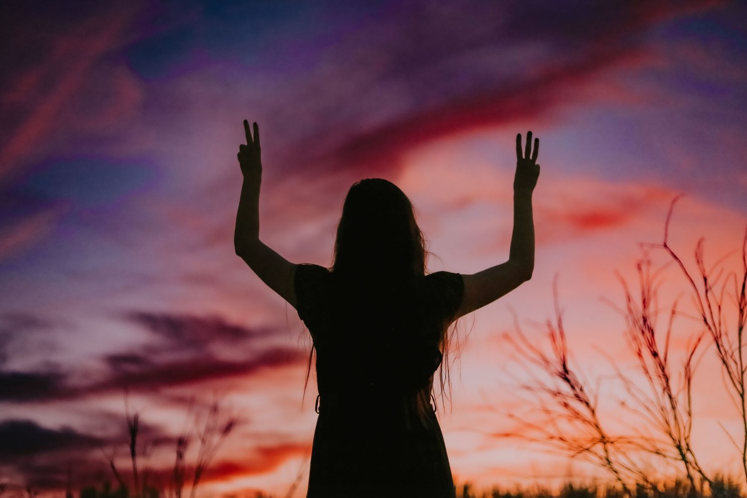 Silhouette of figure making peace signs against dramatic red sunset sky.