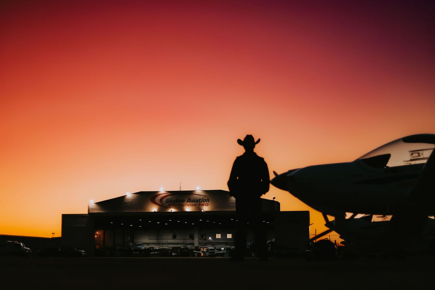Cowboy silhouette standing by truck against orange sunset.