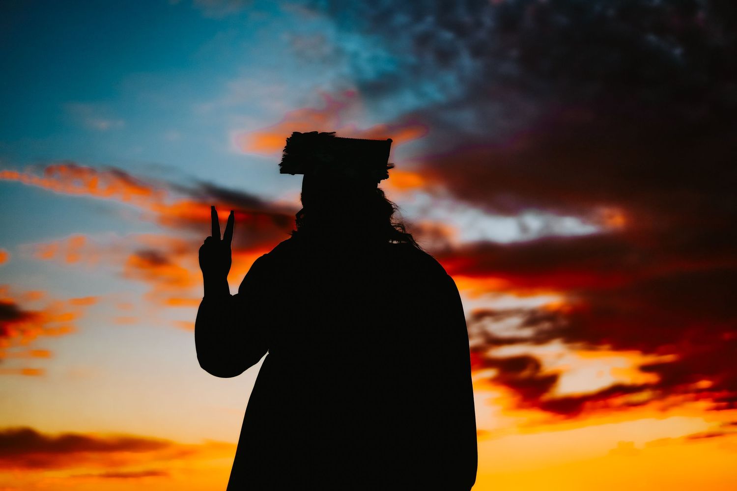 Graduate silhouette making peace sign against colorful sunset sky.