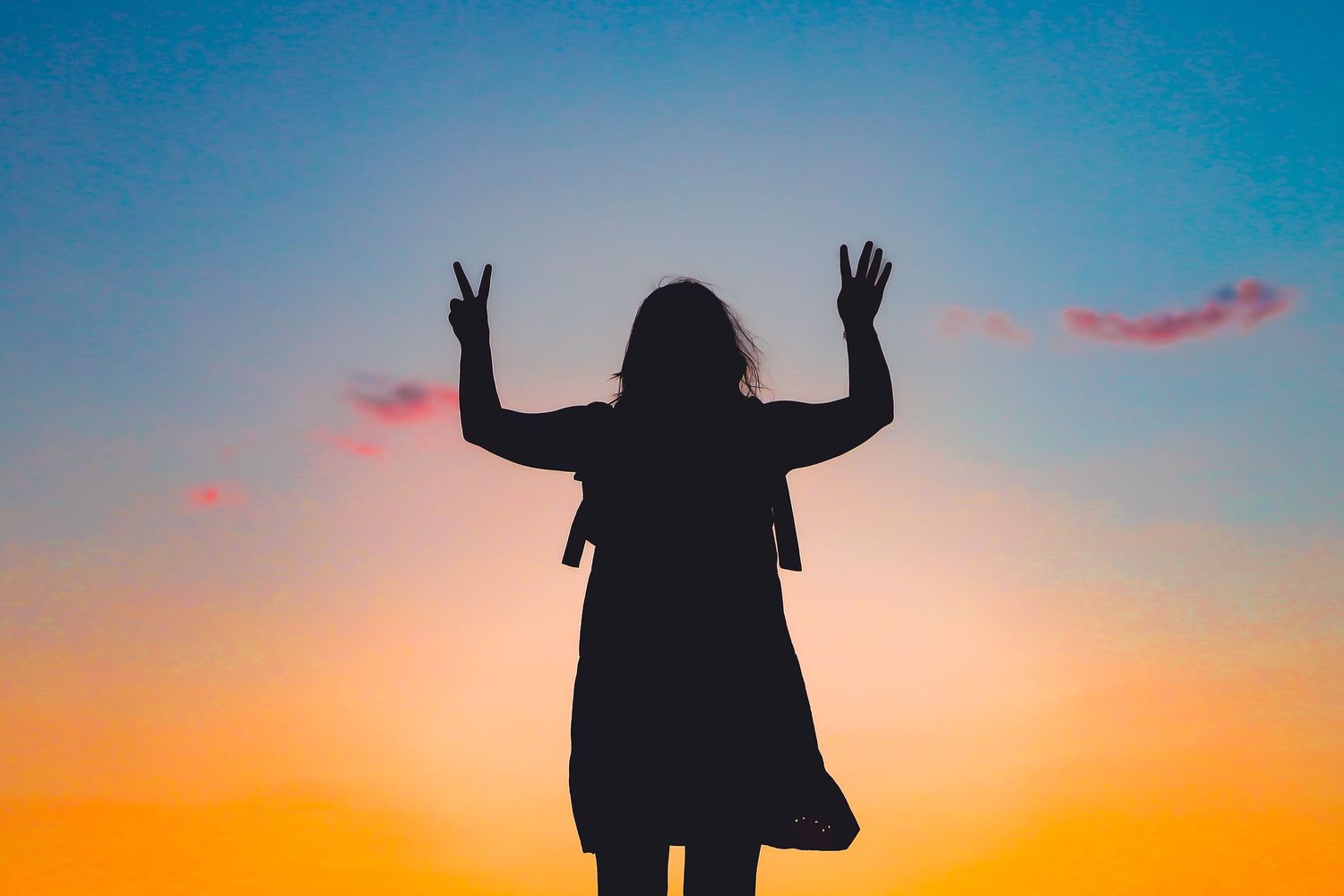 Silhouette of a person raising both hands against a vibrant orange and blue sunset sky.