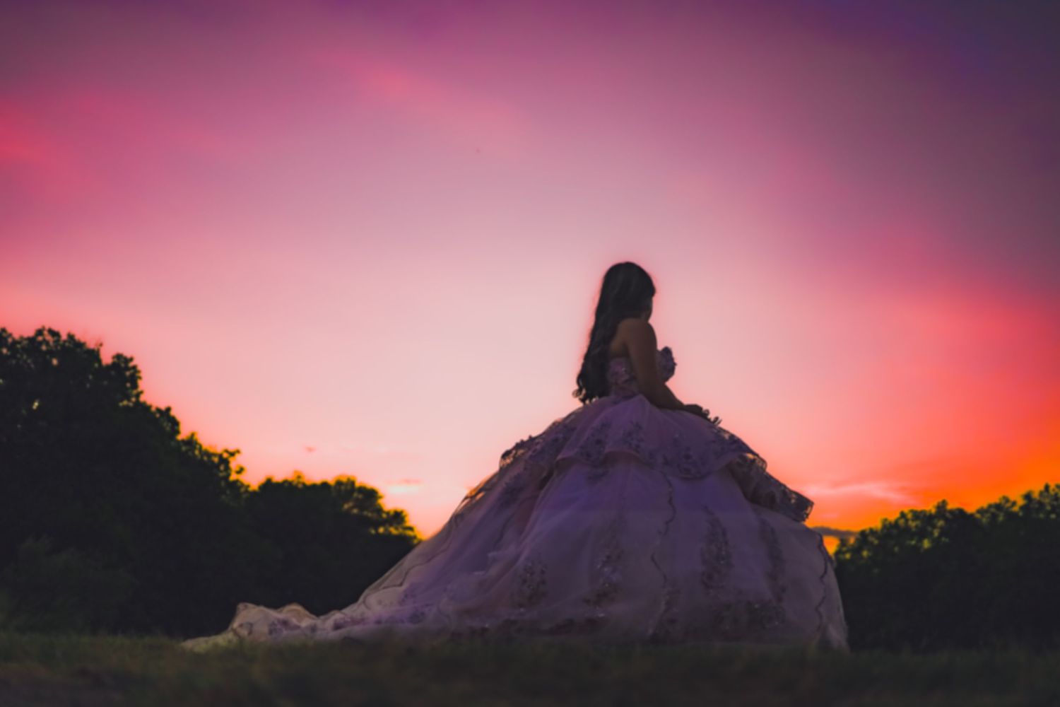 Silhouette of someone in an elaborate dress sitting on a hill during a pink sunset.
