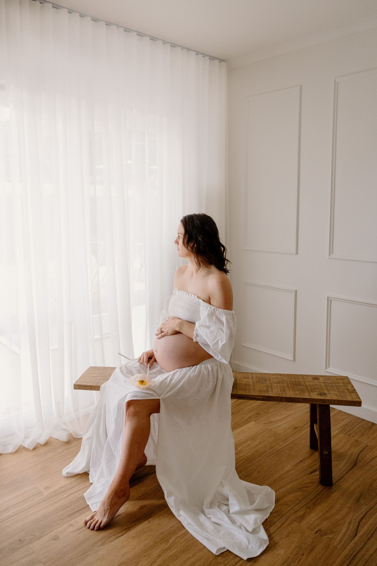 Artistic maternity photo of woman in white dress sitting on wooden bench by window.