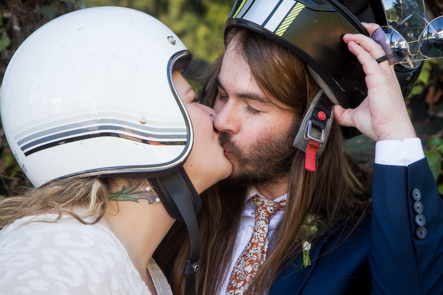 Two people kiss while one wears a retro white motorcycle helmet and the other lifts their own helmet.