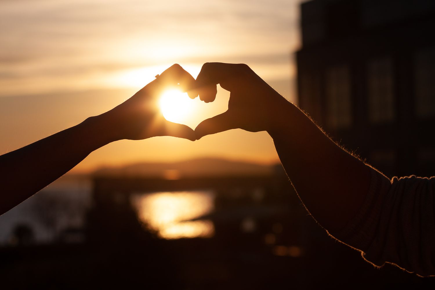 Silhouetted hands form a heart shape against a golden sunset sky with clouds in the background.