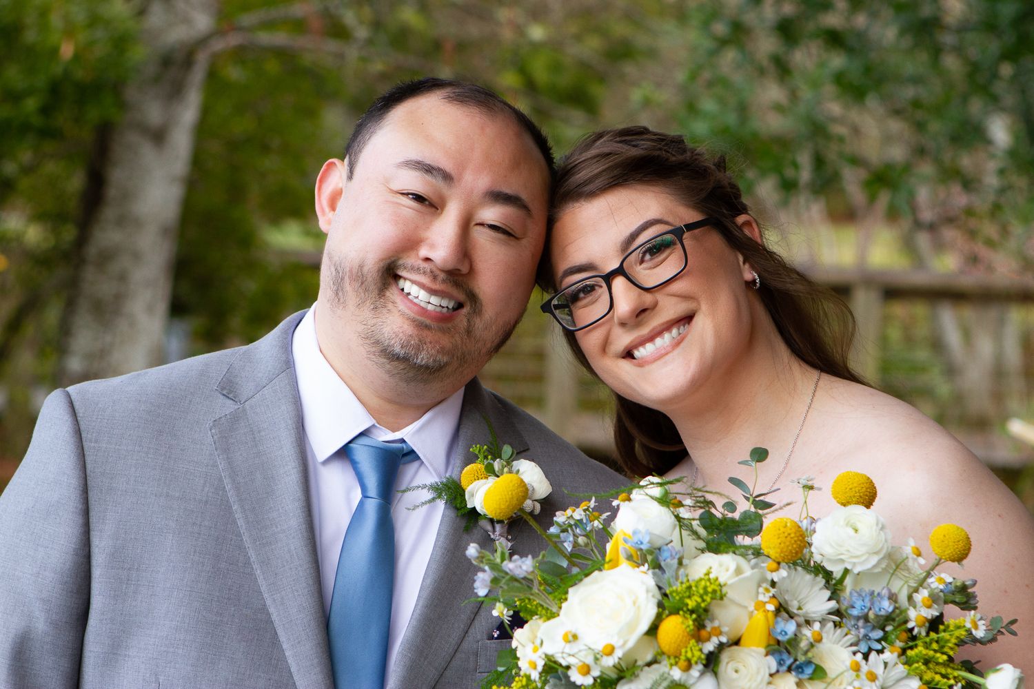 Smiling couple in wedding attire holding yellow and white floral bouquet outdoors on their special day.