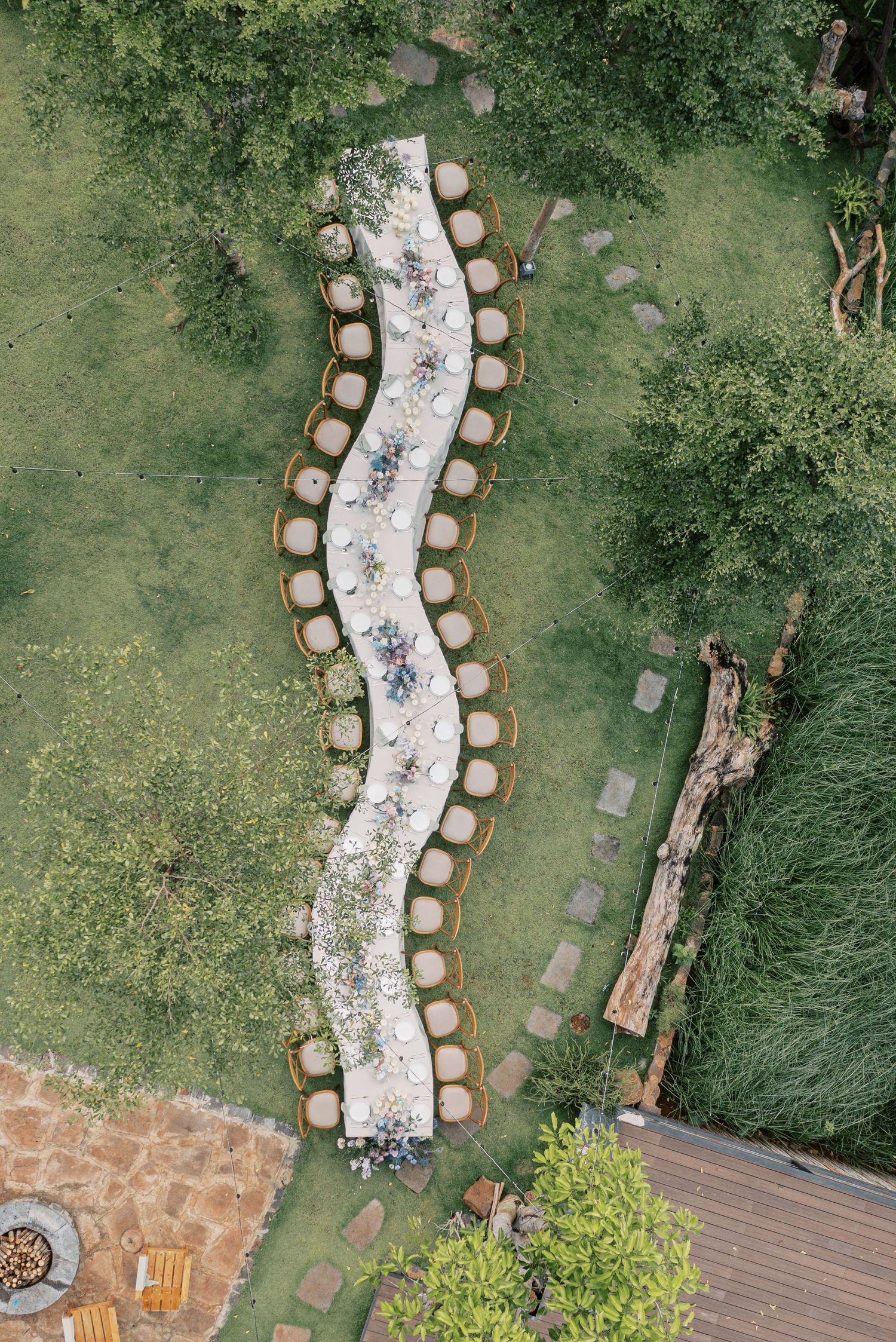 Aerial view of a serpentine outdoor wedding table setting with cream chairs on a lush green lawn.