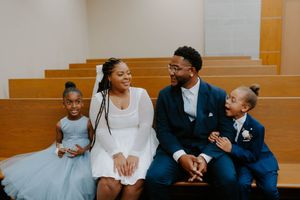 A family in formal attire sits together on wooden church benches, sharing smiles and laughter.