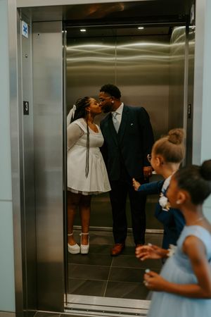 A couple steals a kiss in an elevator while flower girls peek through the doors.