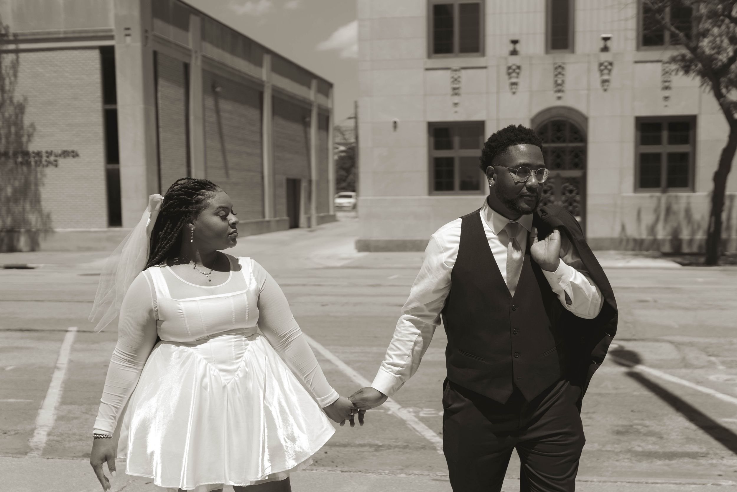 A couple in wedding attire poses together against a white building exterior.