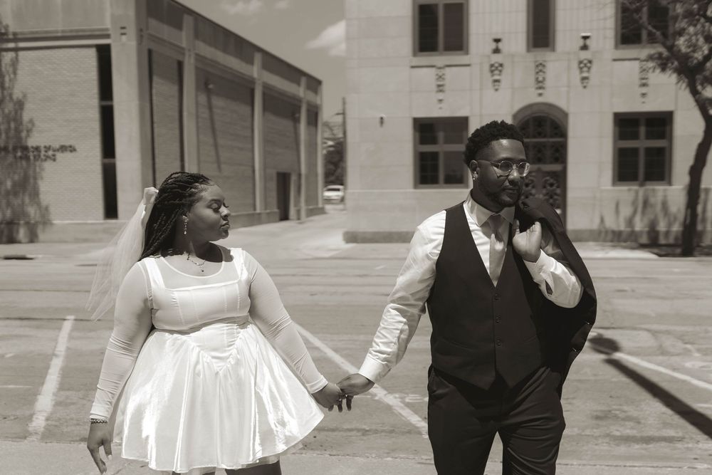 A couple in wedding attire poses together against a white building exterior.