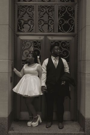 A couple stands in formal attire near an ornate doorway entrance.
