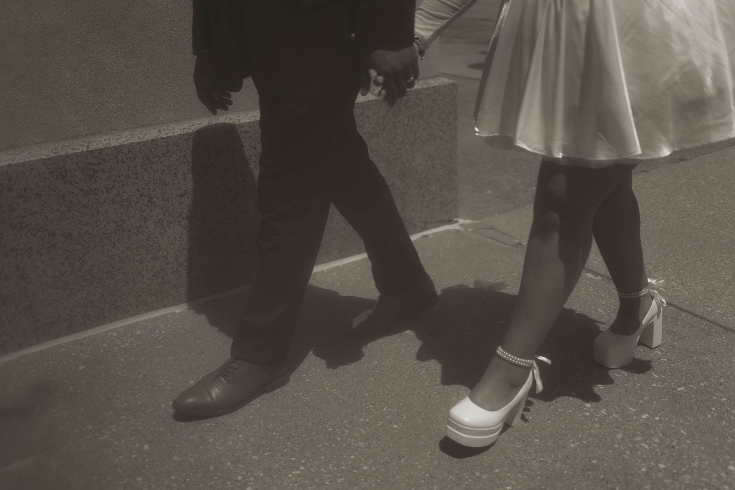 Close-up view of formal shoes and dress hemlines on pavement.