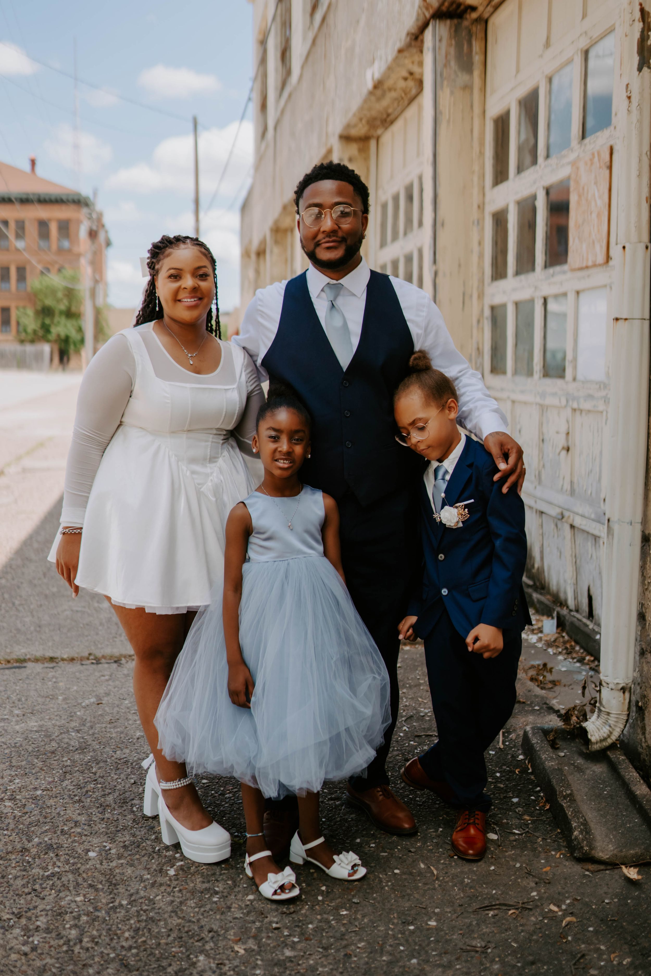 A family poses together against a light stone building wall wearing coordinated white and navy formal attire.