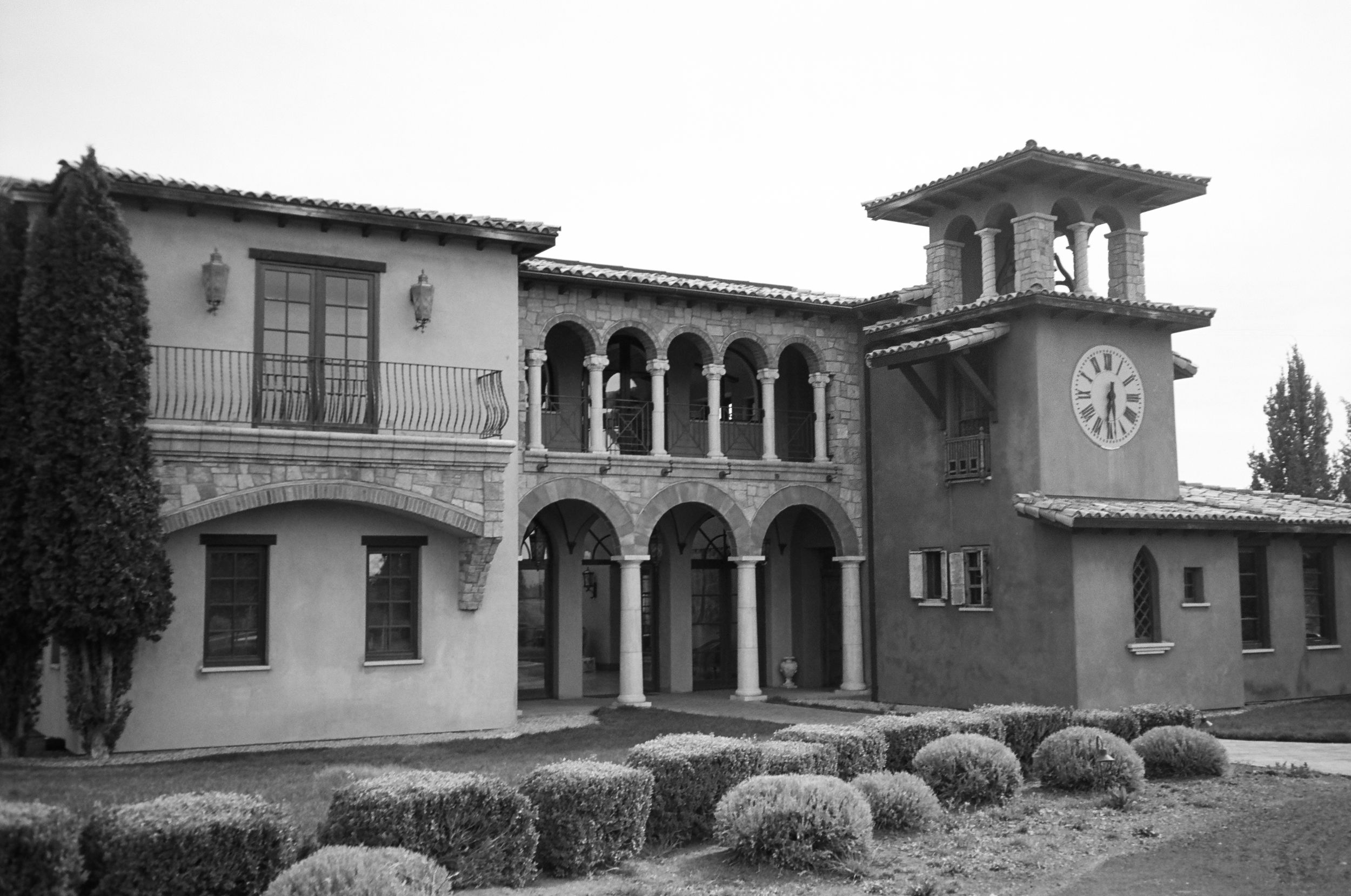 Black and white photo of a Spanish Colonial Revival building with arched colonnade and clock tower surrounded by trimmed hedges.