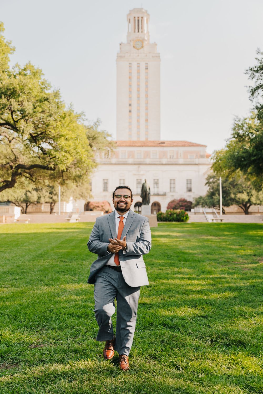A person wearing a gray suit poses on a sunny lawn with a tower visible in the background at UT Austin campus.