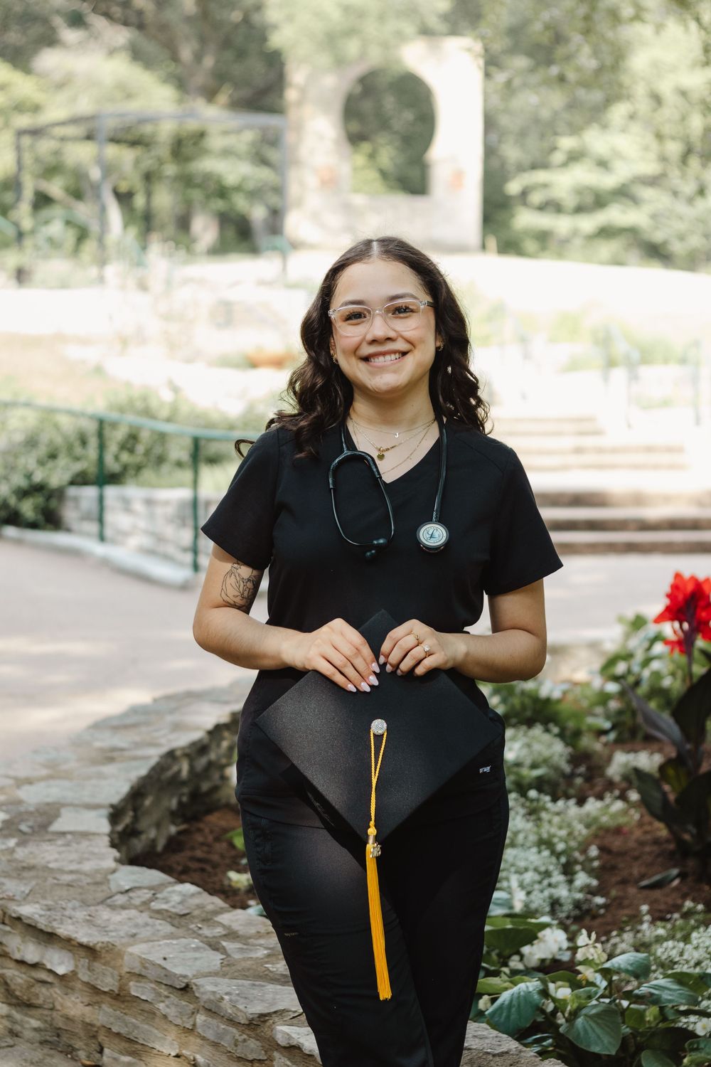 Healthcare professional in black scrubs with stethoscope standing on stone path in garden setting.